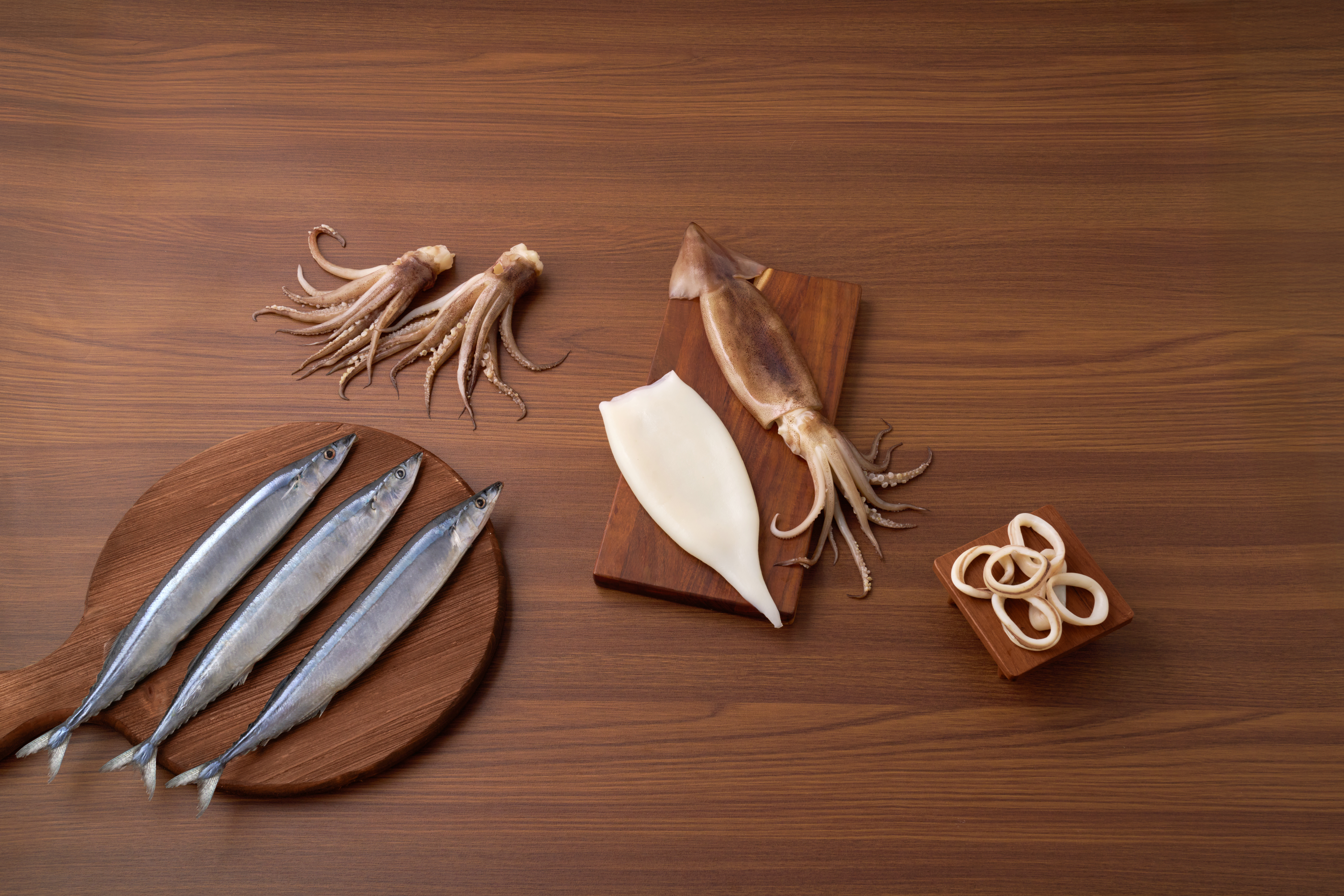 Primary processing — overhead studio shot of graded raw material: saury on a round wooden board, squid (cleaned tube and tentacles) on a rectangular board, and a small dish of squid rings, arranged on dark walnut.